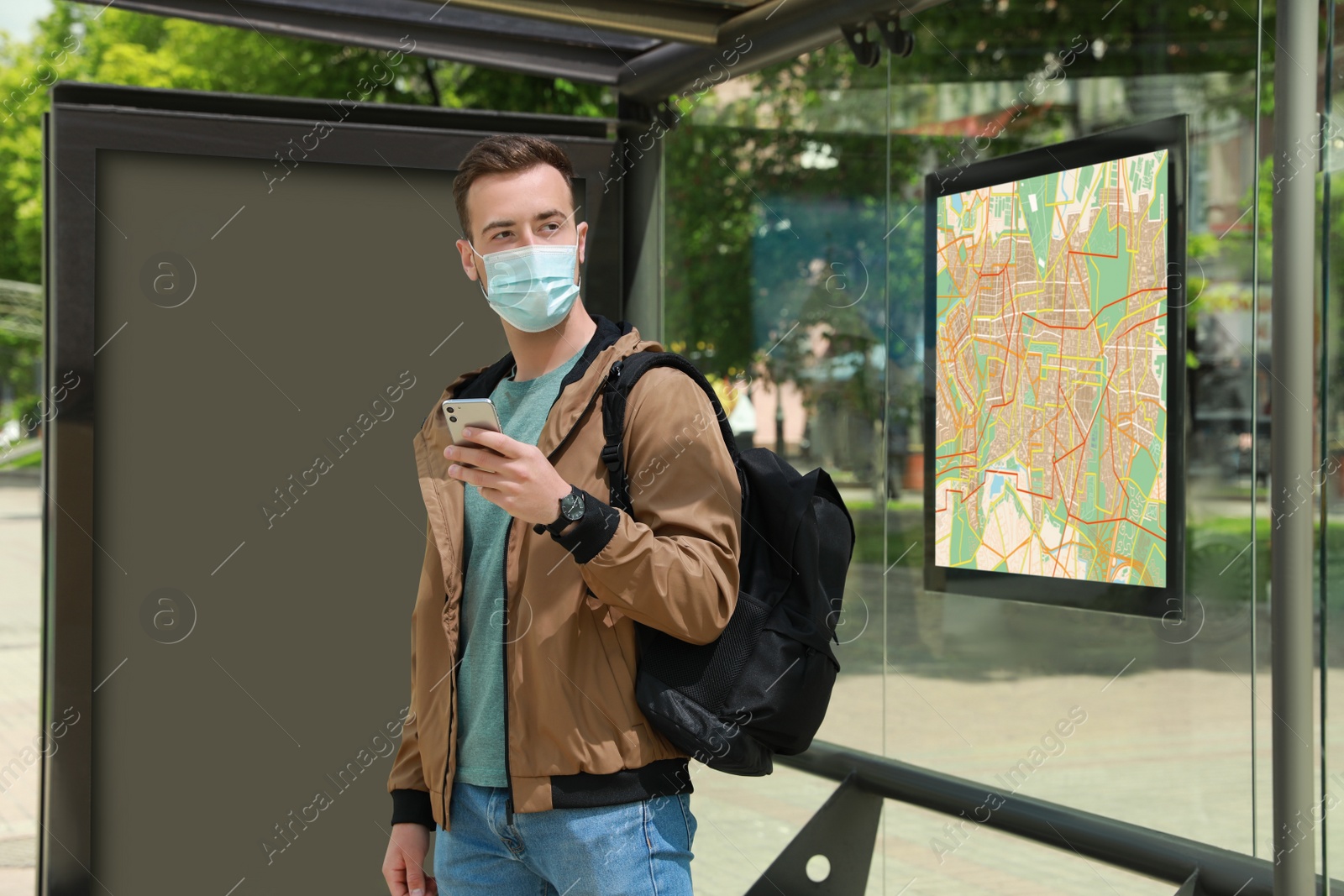 Image of Young man in protective mask with smartphone and backpack waiting for public transport at bus stop
