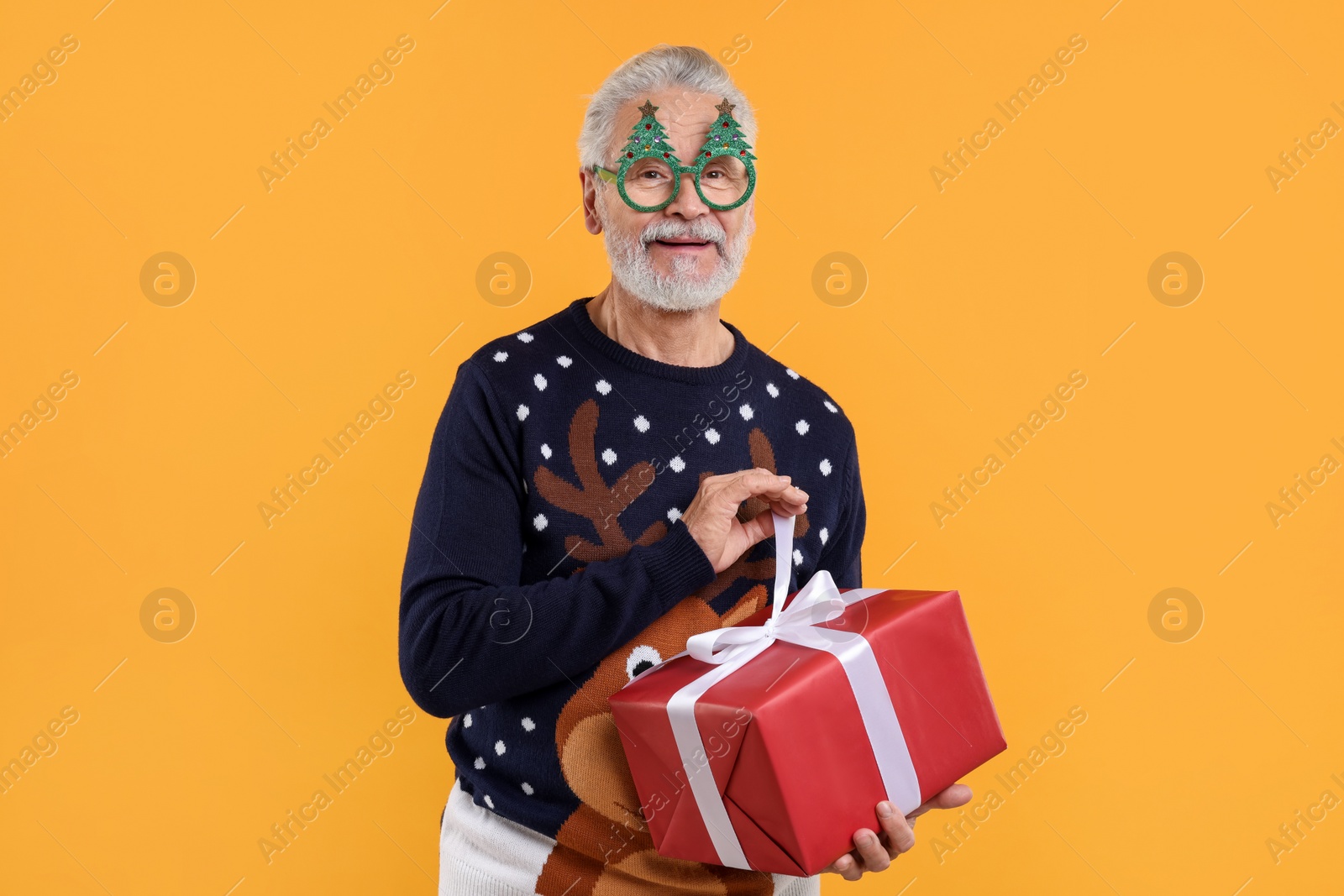 Photo of Senior man in Christmas sweater and funny glasses opening gift against orange background