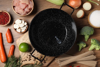 Empty iron wok surrounded by raw ingredients on wooden table, flat lay