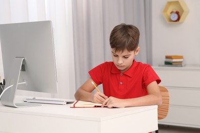 Photo of Boy writing in notepad while using computer at desk in room. Home workplace
