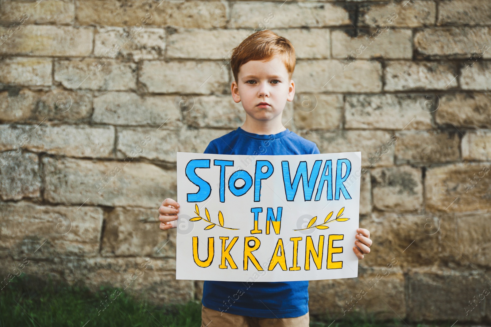 Photo of Sad boy holding poster in colors of national flag with words Stop War In Ukraine against brick wall outdoors