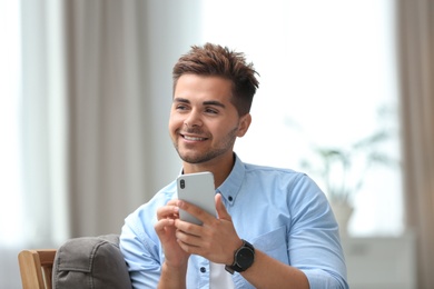 Photo of Portrait of handsome young man with smartphone sitting on chair in room