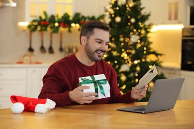Photo of Celebrating Christmas online with exchanged by mail presents. Happy man with greeting card and gift box during video call on laptop at home