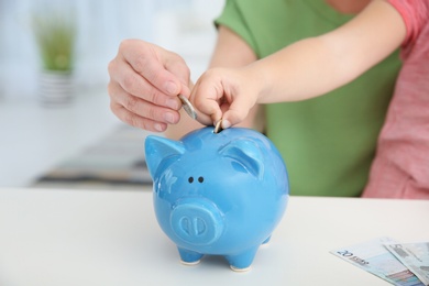 Little girl with grandmother putting money into piggy bank at table, closeup