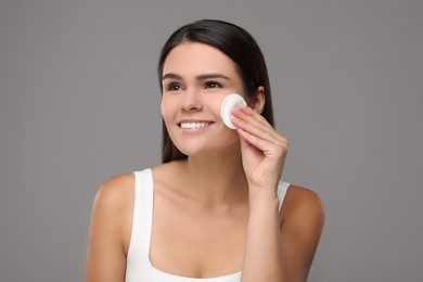 Young woman cleaning her face with cotton pad on grey background