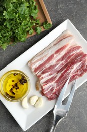 Photo of Pieces of raw pork belly, oil, parsley and garlic on grey textured table, top view