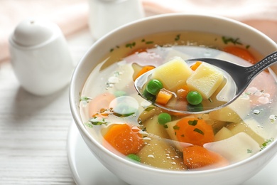 Photo of Spoon of fresh homemade vegetable soup over full bowl on white wooden table, closeup