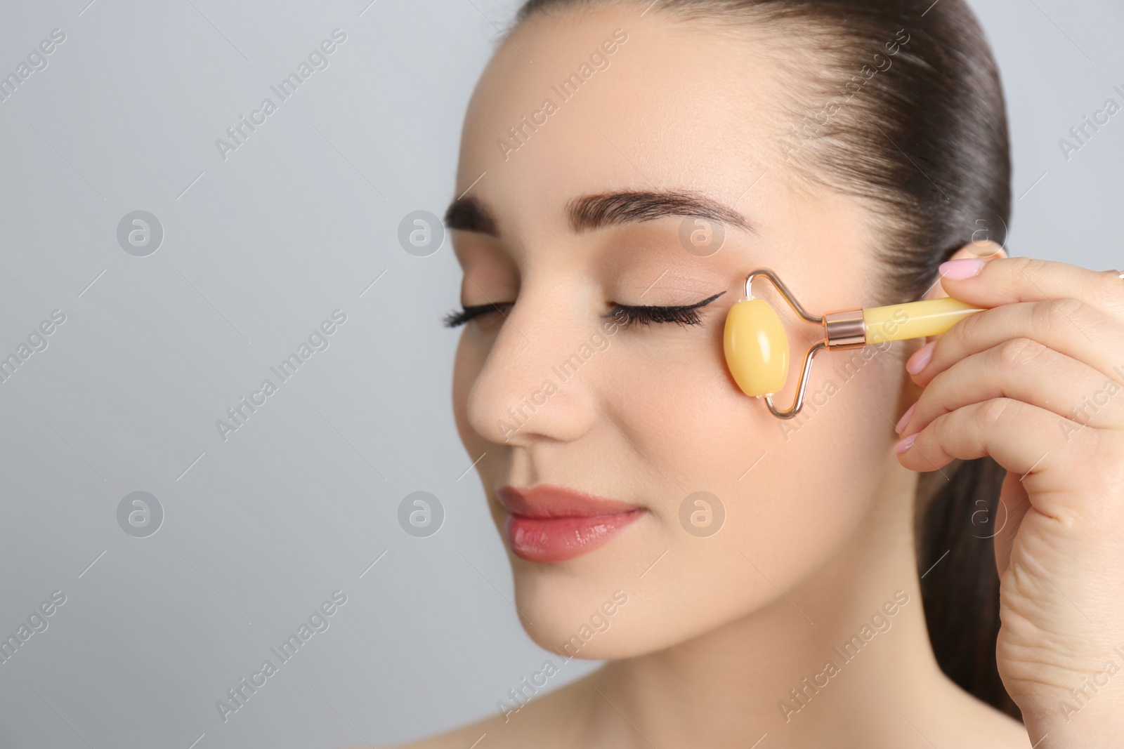 Photo of Woman using natural jade face roller on grey background, closeup. Space for text