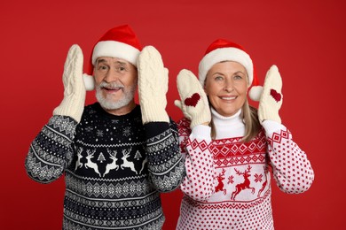 Photo of Senior couple in Christmas sweaters, Santa hats and knitted mittens on red background