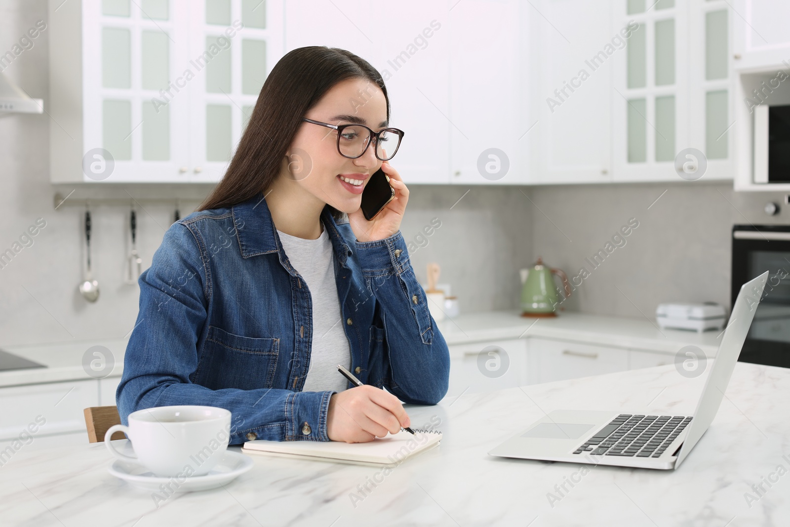 Photo of Home workplace. Happy woman with pen and notebook talking on smartphone at marble desk in kitchen