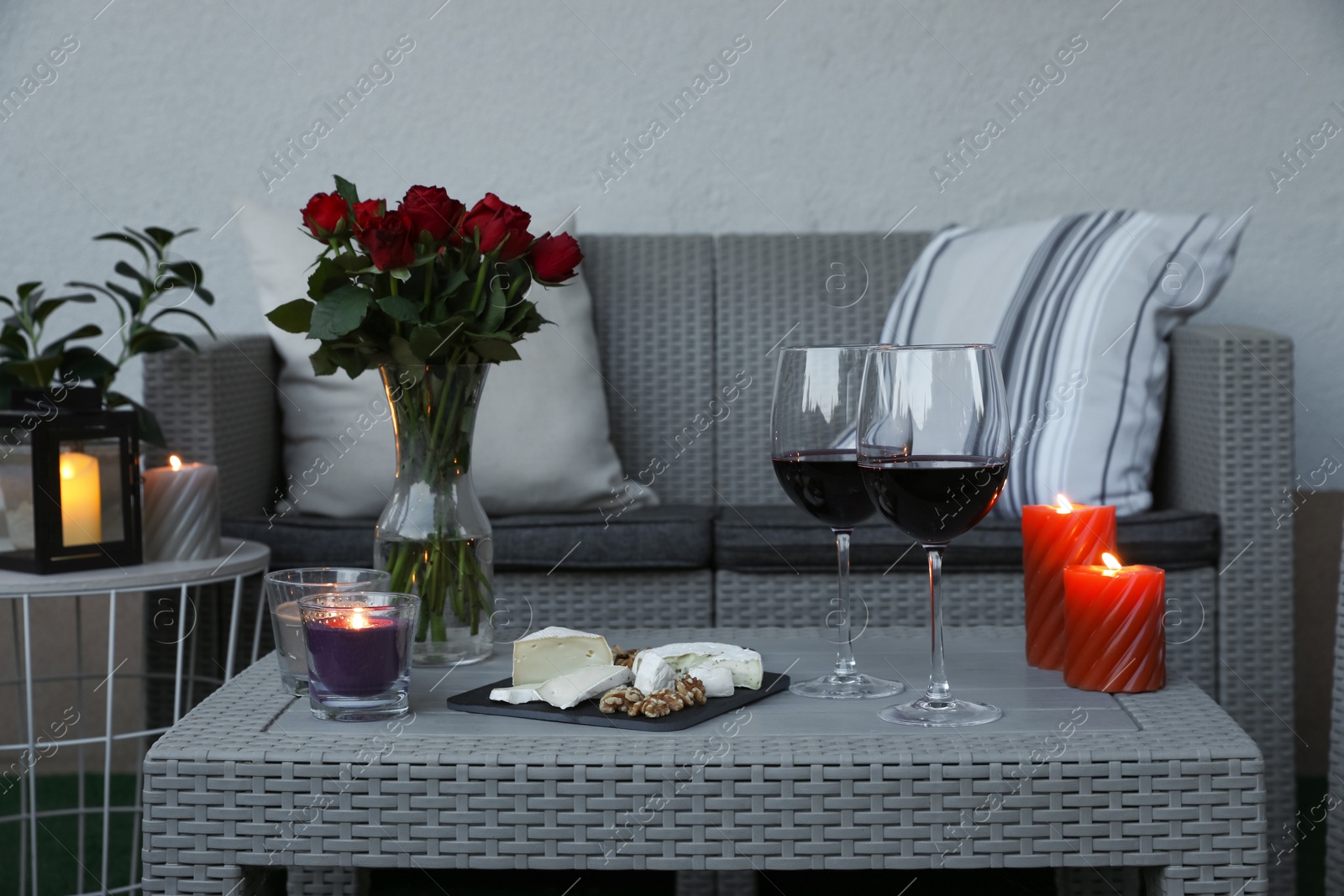 Photo of Glasses of wine, vase with roses, burning candles and snacks on rattan table at balcony in evening