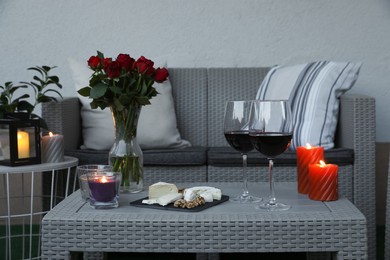 Glasses of wine, vase with roses, burning candles and snacks on rattan table at balcony in evening