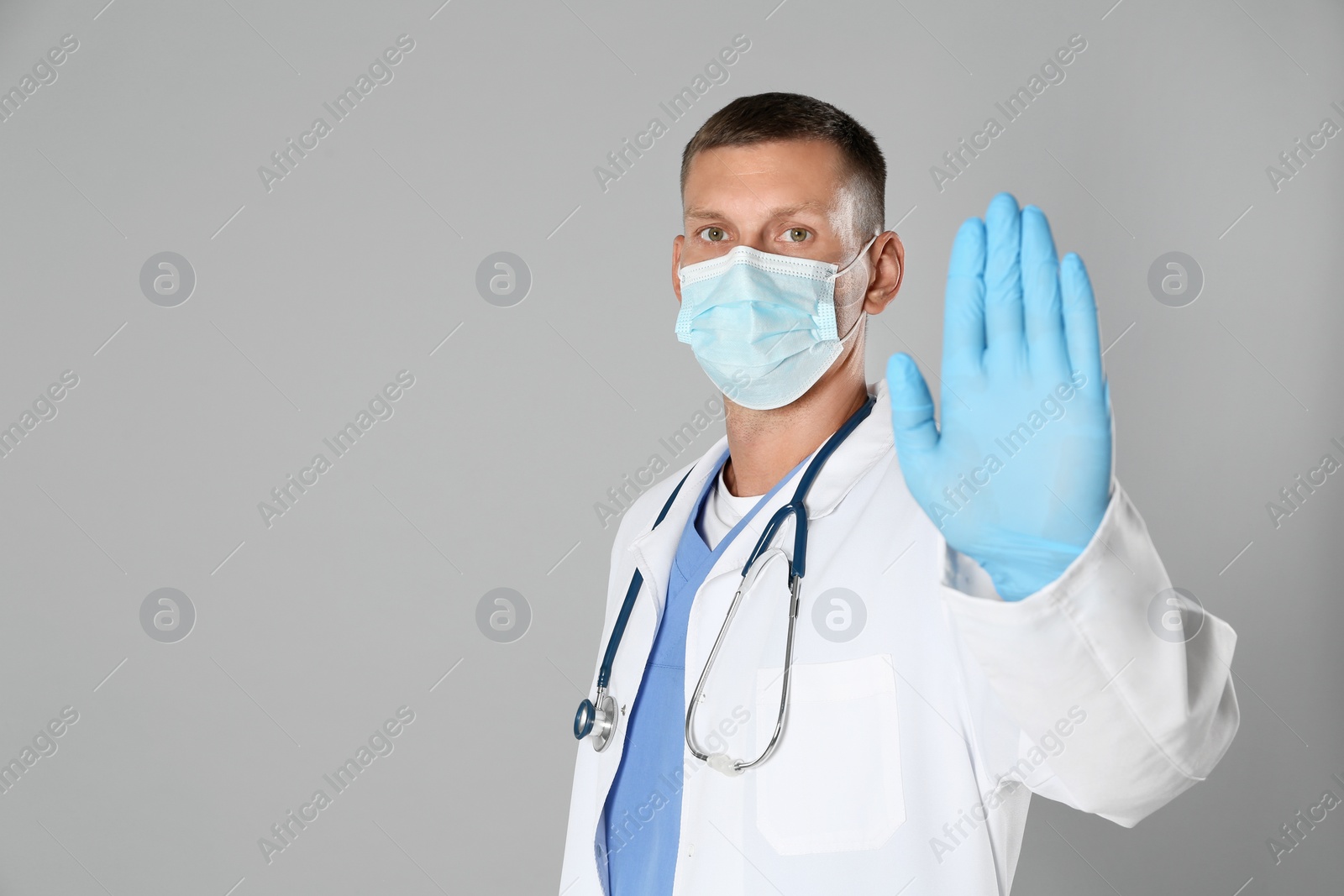 Photo of Doctor in protective mask showing stop gesture on grey background, space for text. Prevent spreading of coronavirus