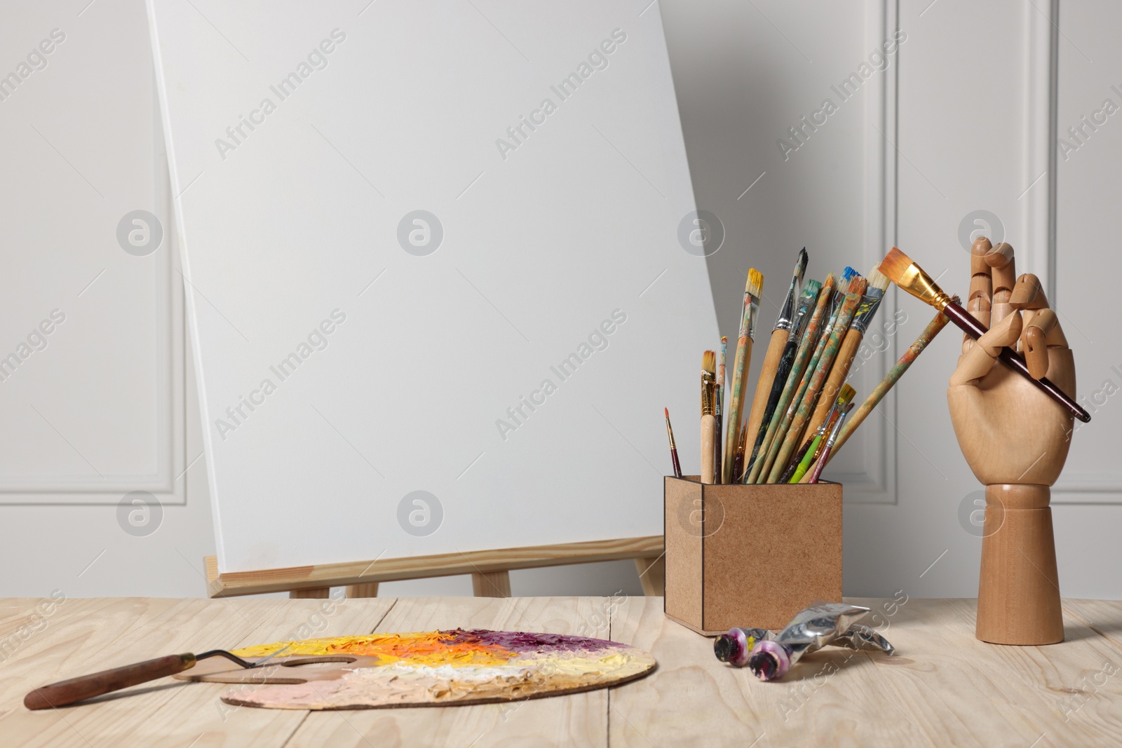 Photo of Easel with blank canvas, hand model and different art supplies on wooden table near white wall