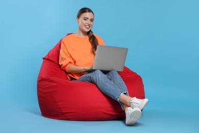 Happy woman with laptop sitting on beanbag chair against light blue background