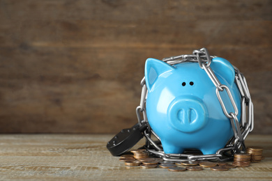 Piggy bank with steel chain, padlock and coins on wooden table, space for text. Money safety concept