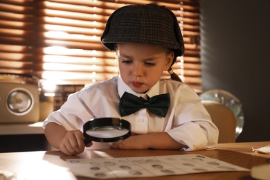 Photo of Cute little detective exploring fingerprints with magnifying glass at table in office