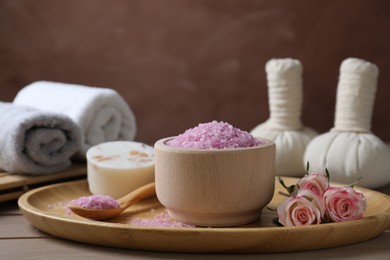 Photo of Bowl of pink sea salt, roses and herbal massage bags on wooden table