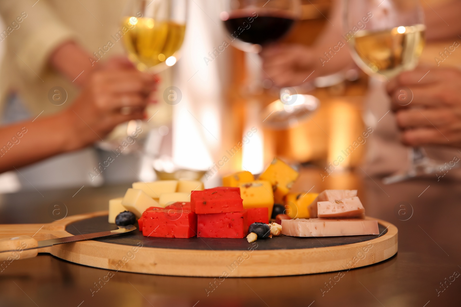 Photo of Different delicious cheeses on table and blurred friends toasting with wine on background