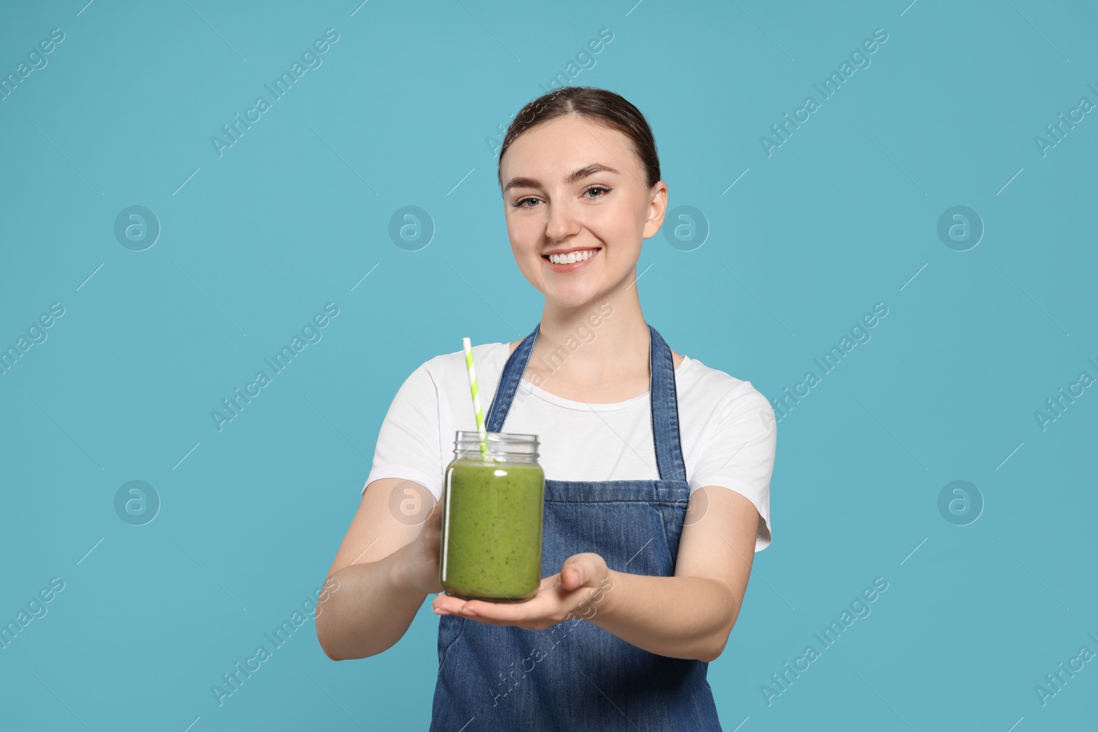 Photo of Beautiful young woman in denim apron with delicious smoothie on light blue background