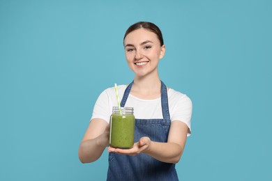 Photo of Beautiful young woman in denim apron with delicious smoothie on light blue background