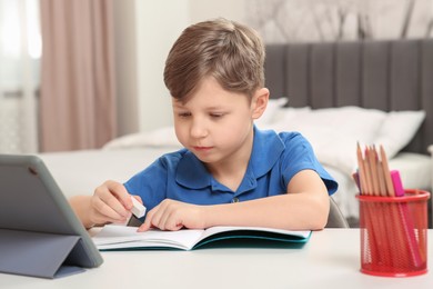 Photo of Little boy erasing mistake in his notebook near tablet at white desk indoors