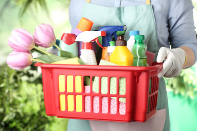 Photo of Woman holding basket with spring flowers and cleaning supplies on green blurred background, closeup
