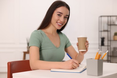 Photo of Young woman with cup of coffee writing in notebook at white table indoors