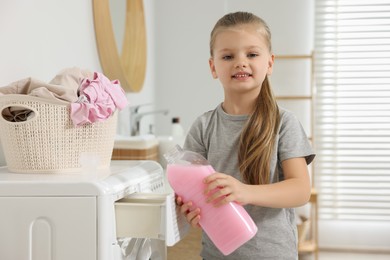 Photo of Little girl pouring fabric softener into washing machine in bathroom