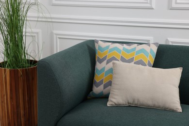 Photo of Green artificial plant in pot and soft sofa with cushions near white wall indoors