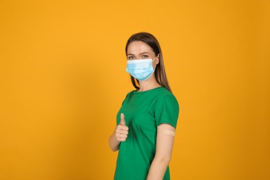 Photo of Vaccinated woman with protective mask and medical plaster on her arm showing thumb up against yellow background