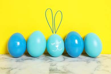 Image of One egg with drawn face and ears as Easter bunny among others on white marble table against yellow background