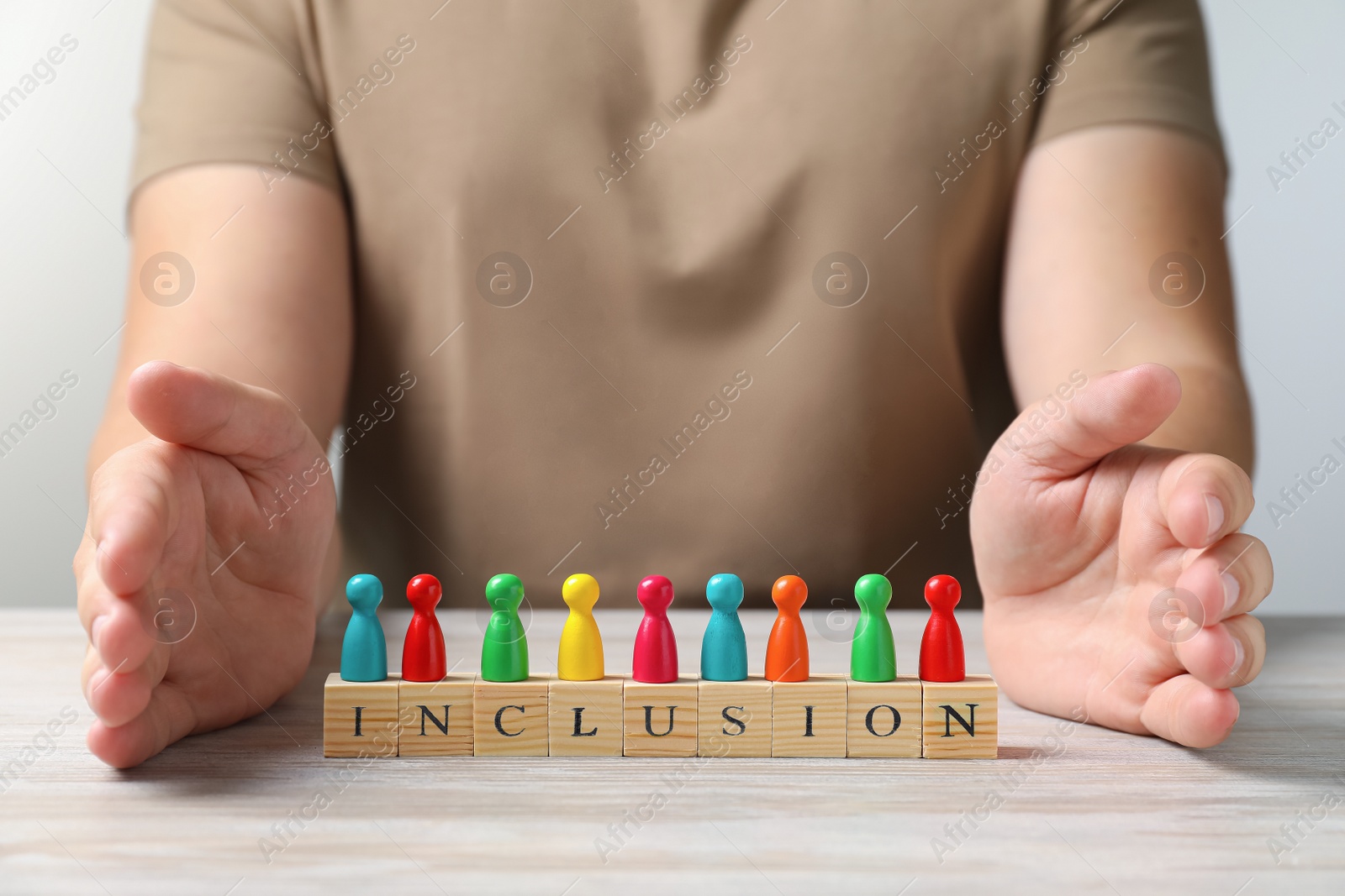 Photo of Woman protecting colorful pawns and cubes with word Inclusion at wooden table, closeup