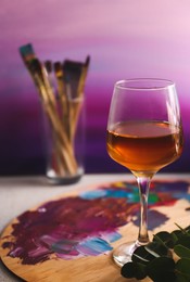 Photo of Wooden artist's palette with colorful paints and glass of wine on table, space for text