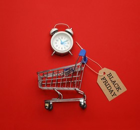 Image of Shopping cart, tag with phrase BLACK FRIDAY and alarm clock on red background, flat lay