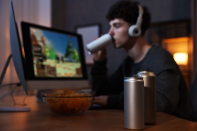 Photo of Young man with energy drink playing video game at wooden desk indoors, focus on can
