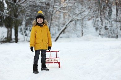 Little boy pulling sledge through snow in winter park, space for text