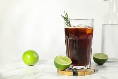 Photo of Refreshing iced coffee with rosemary in glass and fresh limes on white marble table, space for text