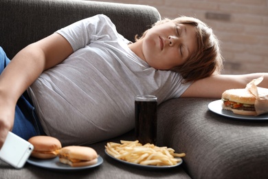 Photo of Overweight boy sleeping on sofa surrounded by fast food