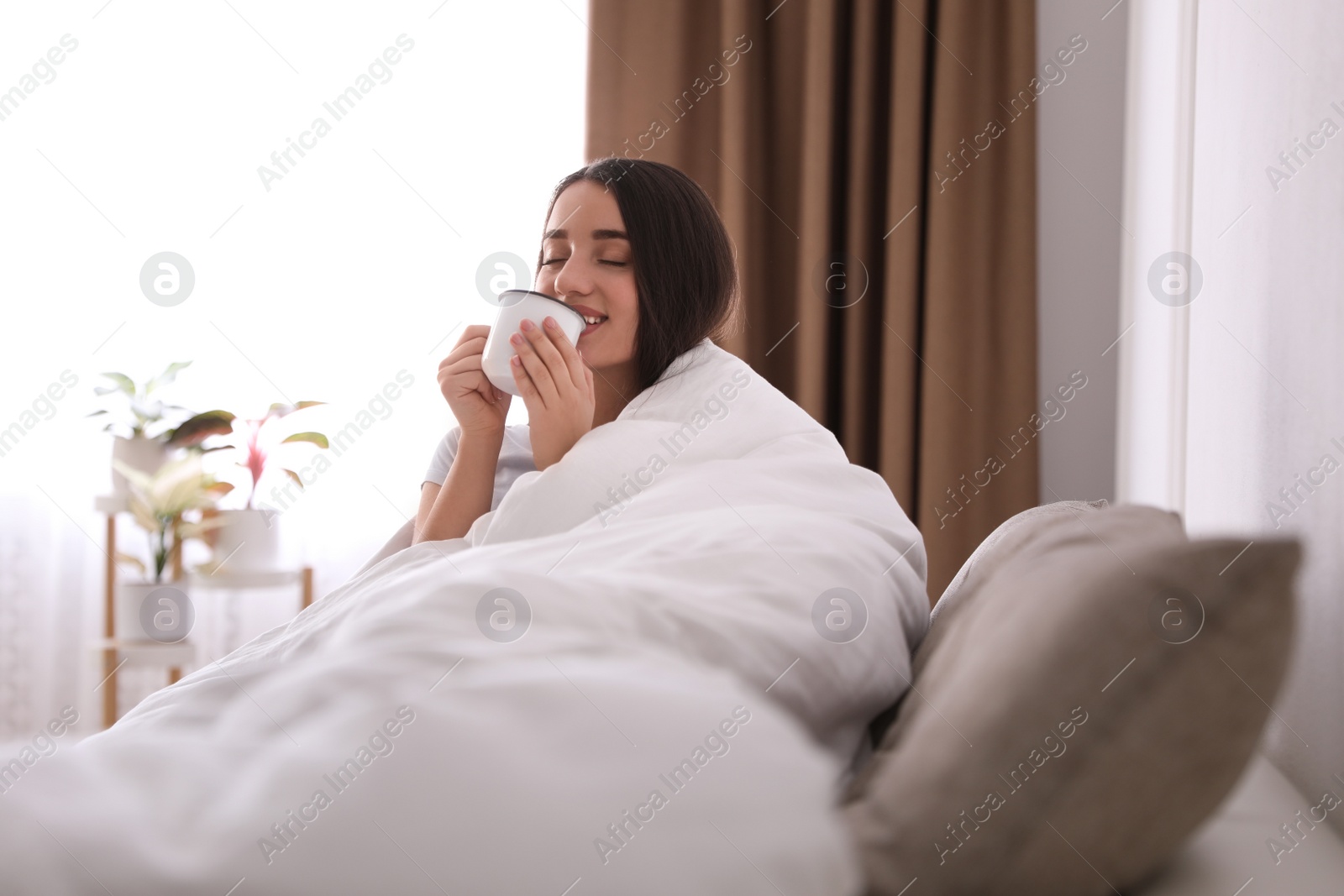 Photo of Woman covered in blanket drinking cup of hot beverage on sofa