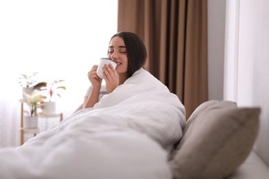 Woman covered in blanket drinking cup of hot beverage on sofa