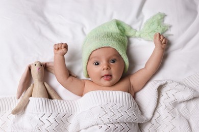 Photo of Cute little baby with toy bunny lying under knitted plaid in bed, top view