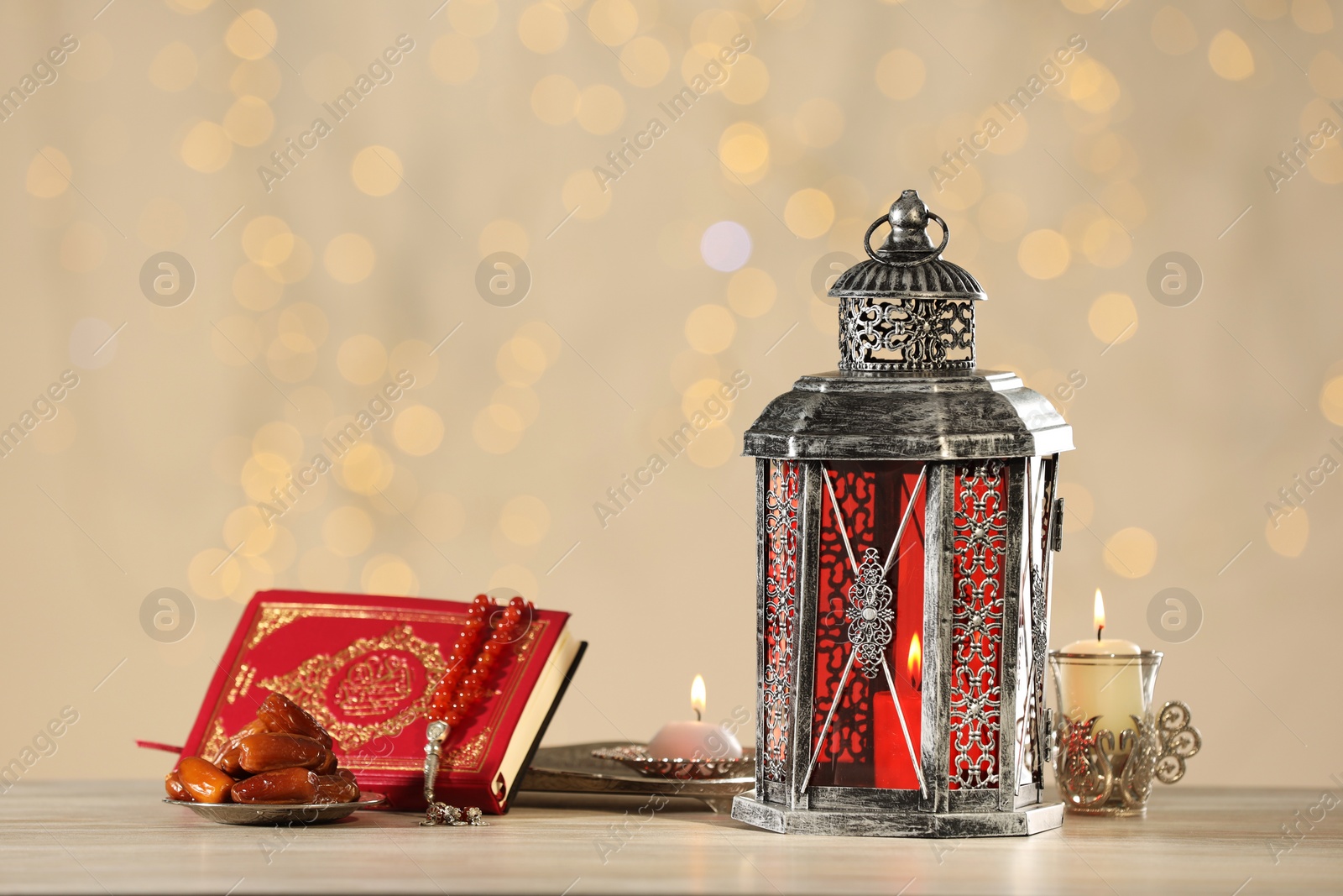 Photo of Arabic lantern, Quran, misbaha, candles and dates on wooden table against blurred lights