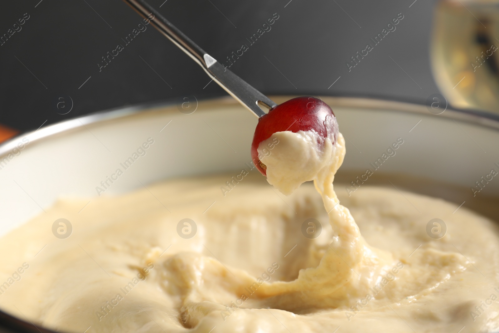 Photo of Dipping piece of grape into fondue pot with melted cheese on grey background, closeup