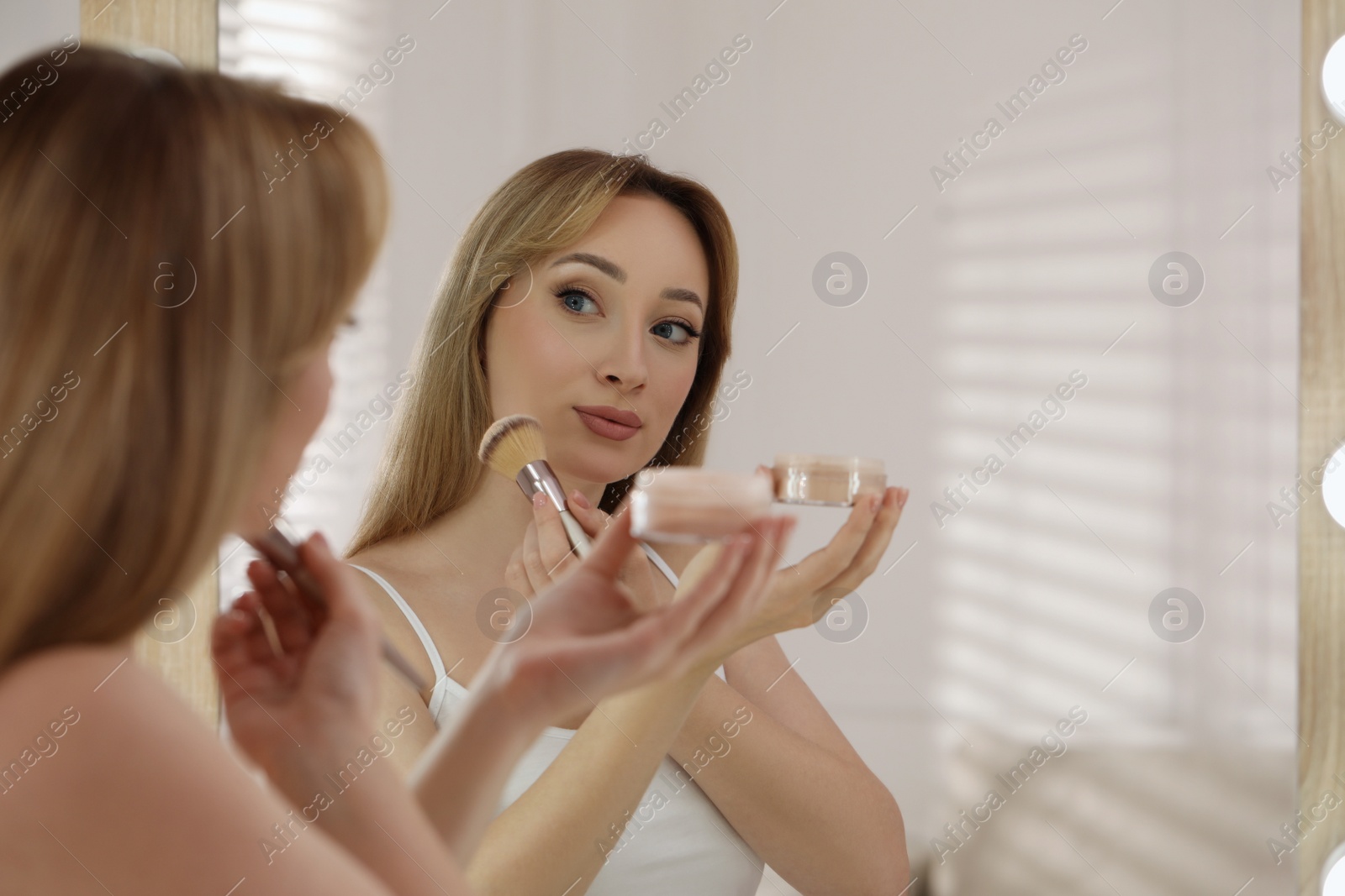 Photo of Beautiful young woman applying face powder with brush in front of mirror at home