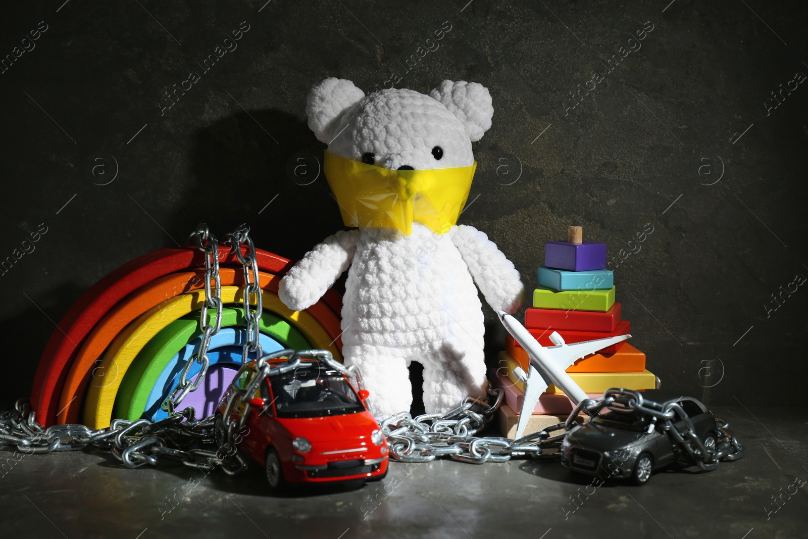 Photo of Stop child abuse. Crochet bear with taped mouth, chain and toys in dark room