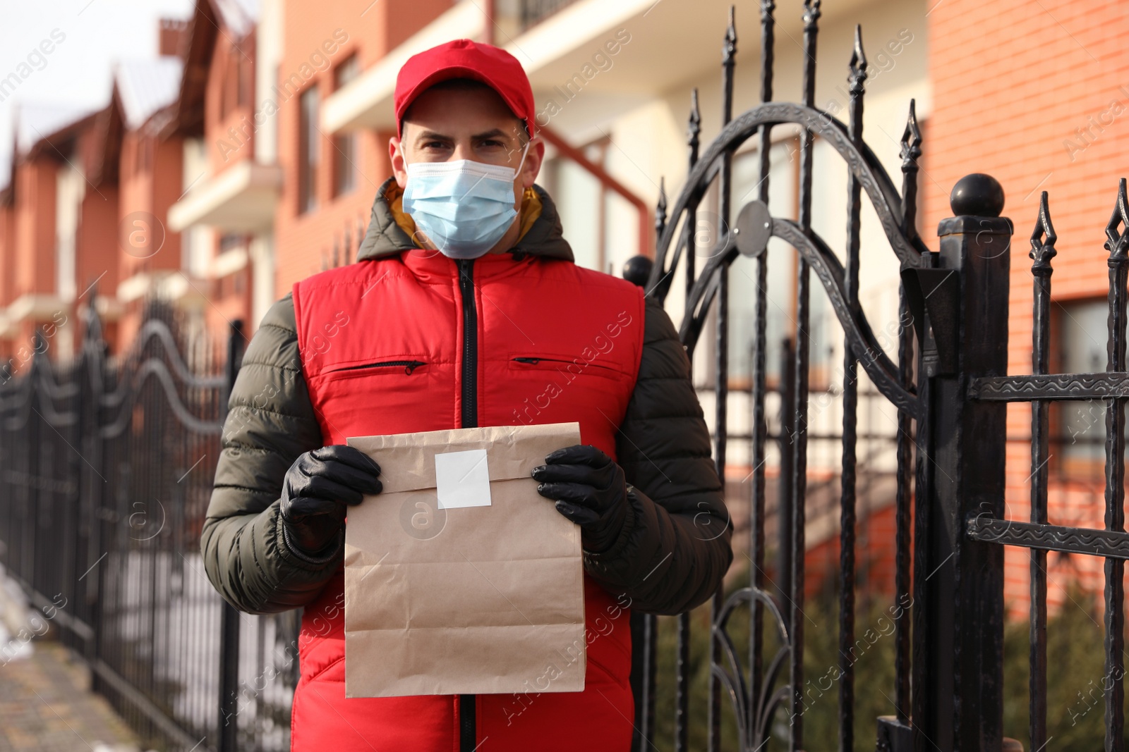 Photo of Courier in medical mask holding paper bag with takeaway food near house outdoors. Delivery service during quarantine due to Covid-19 outbreak