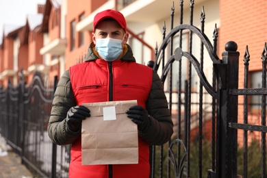 Courier in medical mask holding paper bag with takeaway food near house outdoors. Delivery service during quarantine due to Covid-19 outbreak