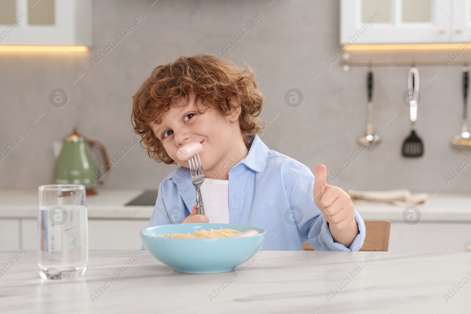 Photo of Cute little boy eating sausage and showing thumbs up at table in kitchen