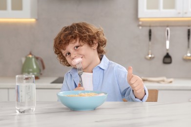 Cute little boy eating sausage and showing thumbs up at table in kitchen